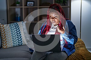 African american woman with braided hair using computer laptop at night in shock face, looking skeptical and sarcastic, surprised