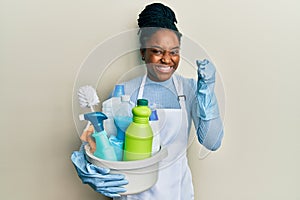 African american woman with braided hair wearing apron holding cleaning products screaming proud, celebrating victory and success