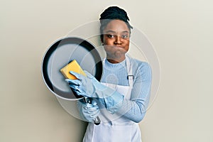 African american woman with braided hair wearing apron holding scourer washing pan puffing cheeks with funny face