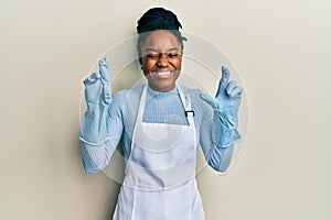 African american woman with braided hair wearing cleaner apron and gloves gesturing finger crossed smiling with hope and eyes