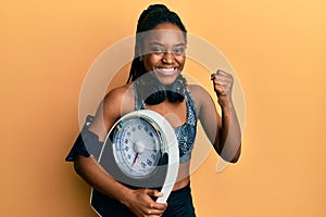 African american woman with braided hair wearing sportswear holding weighing machine screaming proud, celebrating victory and