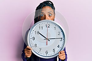 African american woman with braids holding big clock covering face afraid and shocked with surprise and amazed expression, fear