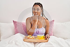 African american woman with braids holding tray with breakfast food in the bed covering mouth with hand, shocked and afraid for