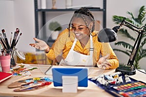 African american woman with braids sitting at art studio painting looking at tablet celebrating achievement with happy smile and