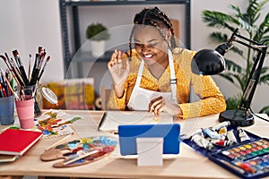 African american woman with braids sitting at art studio painting looking at tablet looking positive and happy standing and