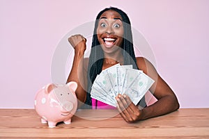 African american woman with braids sitting on the table with piggy bank and dollars screaming proud, celebrating victory and