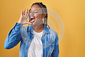 African american woman with braids standing over yellow background shouting and screaming loud to side with hand on mouth