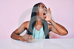 African american woman with braids wearing casual clothes sitting on the table shouting and screaming loud to side with hand on