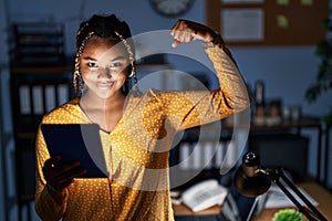 African american woman with braids working at the office at night with tablet strong person showing arm muscle, confident and