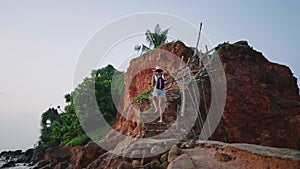 African american woman climbing stairs with backpack on tropical rocky island. Multiracial female in straw hat hikes by
