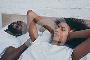 African american woman covering ears with hands while lying near snoring husband