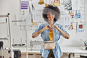 African-American woman designer shows heart with hands near sewing machine in workshop
