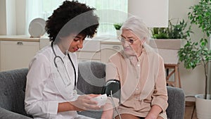 African american woman doctor checking blood pressure of senior woman while sitting on couch at home