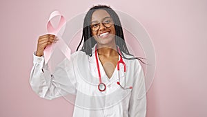 African american woman doctor holding breast cancer awareness pink ribbon over isolated pink background