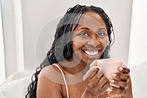 African american woman drinking cup of coffee sitting on bed at bedroom