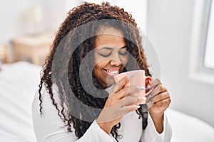 African american woman drinking cup of coffee sitting on bed at bedroom