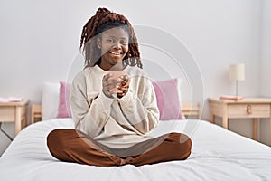 African american woman drinking cup of coffee sitting on bed at bedroom