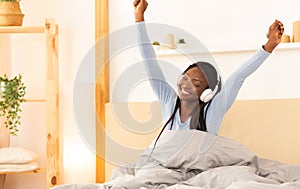 African American Woman In Headphones Stretching Arms Sitting In Bed