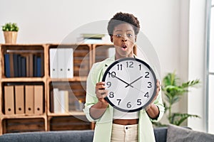 African american woman holding big clock afraid and shocked with surprise and amazed expression, fear and excited face