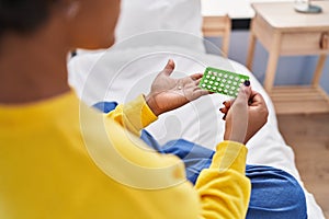 African american woman holding birth control pills sitting on bed at bedroom