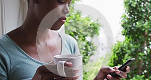 African american woman holding coffee cup using smartphone at home