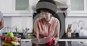 African american woman holding coffee cup using smartphone in the kitchen at home