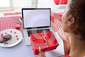 African american woman holding gift box blowing a kiss on videocall on laptop at home