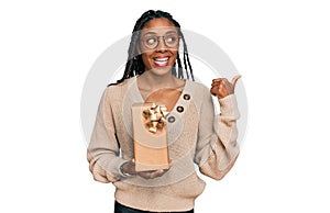 African american woman holding gift pointing thumb up to the side smiling happy with open mouth