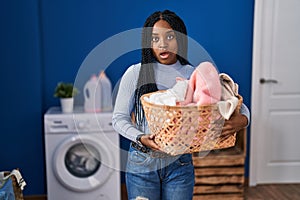 African american woman holding laundry basket scared and amazed with open mouth for surprise, disbelief face