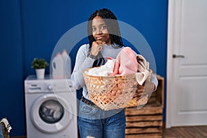 African american woman holding laundry basket serious face thinking about question with hand on chin, thoughtful about confusing