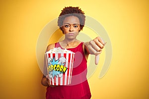 African american woman holding pack of popcorn over yellow isolated background with angry face, negative sign showing dislike with