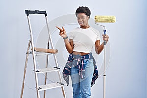 African american woman holding roller painter with a big smile on face, pointing with hand finger to the side looking at the