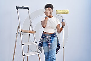 African american woman holding roller painter covering one eye with hand, confident smile on face and surprise emotion