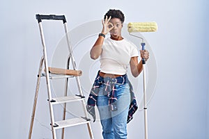 African american woman holding roller painter doing ok gesture shocked with surprised face, eye looking through fingers