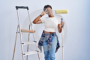 African american woman holding roller painter doing peace symbol with fingers over face, smiling cheerful showing victory