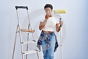 African american woman holding roller painter laughing and embarrassed giggle covering mouth with hands, gossip and scandal