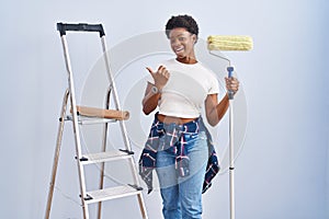 African american woman holding roller painter pointing to the back behind with hand and thumbs up, smiling confident