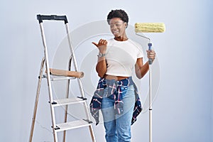 African american woman holding roller painter smiling with happy face looking and pointing to the side with thumb up