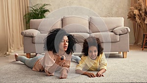 African American woman and litle girl watching TV switching channels with remote control. Mother and daughter lying on