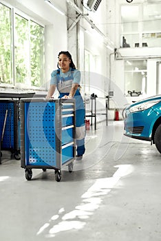 African american woman, professional female mechanic in uniform pulling, carrying tool box cart in auto repair shop. Car