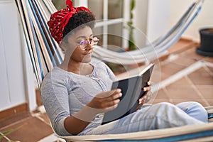 African american woman reading book lying on hammock at home terrace