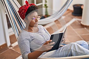 African american woman reading book lying on hammock at home terrace