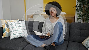 African american woman reading book sitting on sofa at home