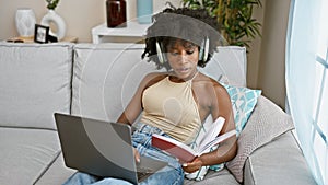 African american woman reading book using laptop and headphones at home