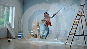 African American woman in red headphones paints wall with white paint using long paint roller and enjoy the music. Black