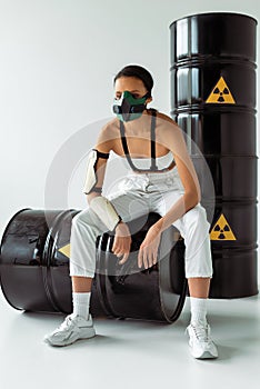 African american woman in safety mask with gun near radioactive waste barrels on white background