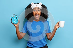 African American woman screaming with clock alarm and coffee in the hand, over blue background