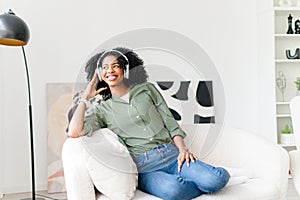 An African-American woman sits on a sofa, smiling while wearing headphones
