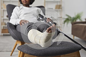 African american woman is sitting at home in armchair with broken bandaged leg lying on stool and holding crutches in