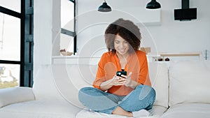 African American woman sitting home couch looking smartphone using phone play mobile game win victory reaction receive